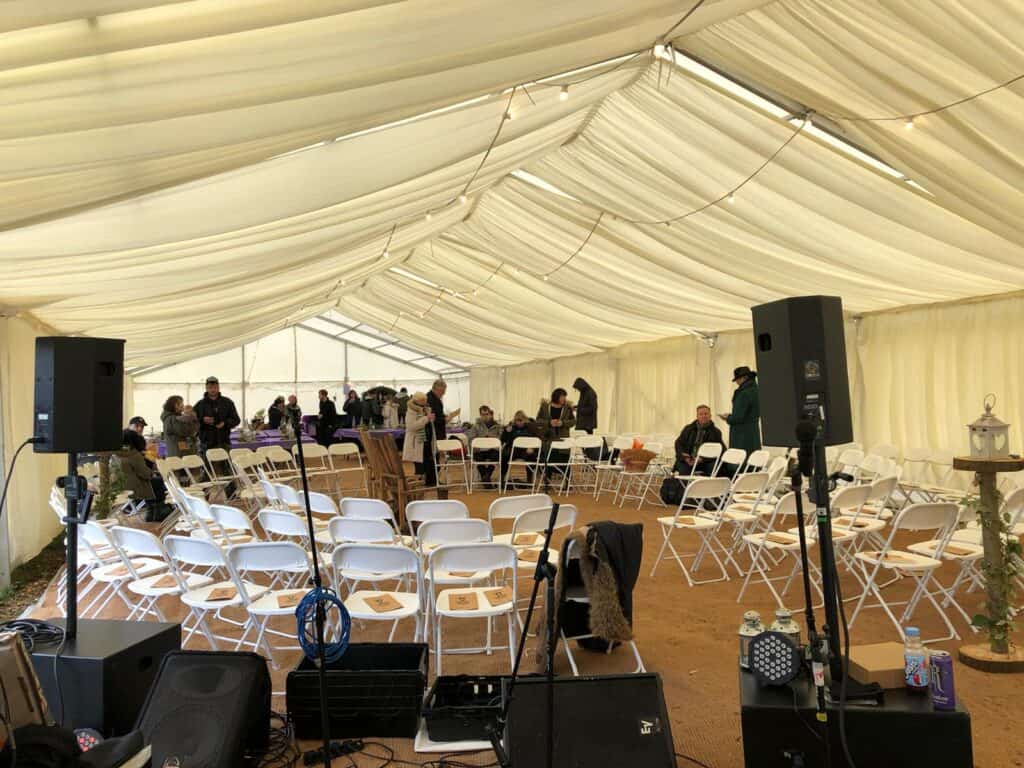 A ceremony inside a clear span modular marquee, with a circle of chairs, a PA setup, flooring, and festoon lights and linings decorating the marquee.